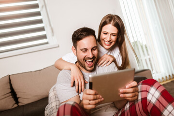 Young couple watching media content online in a tablet sitting on a sofa in the living room.