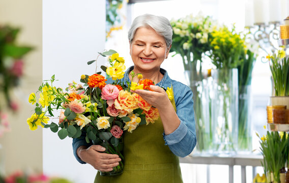 Gardening, Floristics And Old People Concept - Portrait Of Smiling Senior Woman In Green Garden Apron With Bunch Of Flower Over Shop Background