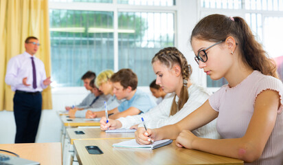 Fototapeta premium Smart teenagers studying in classroom, listening to lecturer and writing in notebooks