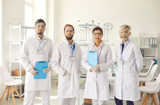 Portrait of medical research group members of different ages in white lab coats, gloves and protective goggles. Team of serious young and mature scientists standing in laboratory and looking at camera