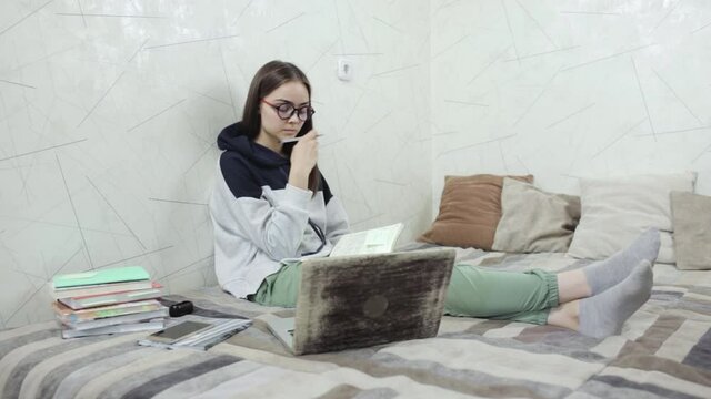 Smart woman preparing for exam at home.  Thoughtful young female student taking notes in notebook and doing homework while sitting on bed and studying online 
