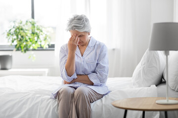 health, old age and people concept - senior woman in pajamas suffering from headache sitting on bed...