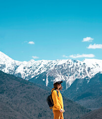 side view Young blond woman in yellow hoodie, cap, with backpack on background of snow capped Caucasus mountains. Hiking and travel, active lifestyle, copy space