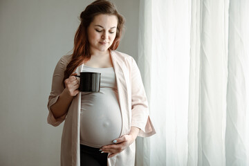 A young pregnant woman with a cup of hot drink stands near the window.