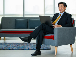 60s Asian senior successful executive business male entrepreneur sitting, leaning back on sofa in the indoor office, using laptop for technology, working with confidence, smiling with positivity.