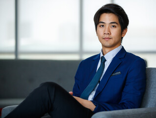 20s visionary young handsome Asian executive businessman wearing a formal suit, sitting cross-legged on comfortable with relaxation, looking at camera with determination and confidence in success.