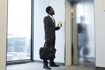 Minimal full length portrait of African-American businessman waiting for elevator in office building and holding coffee cup, copy space © Seventyfour