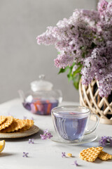 Decorative composition with a Bouquet of lilacs on a light kitchen interior. A cup of purple tea, tea pot and waffles. Bouquet of lilacs in a vase. Spring floral background with lilacs. Tea drinking