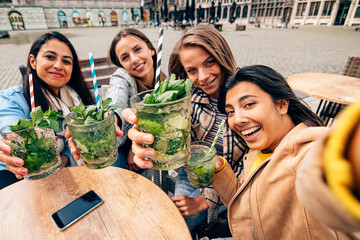 focus on mojito drinks held by a multiracial group of female friends taking a selfie while sitting at cafe table outdoor.