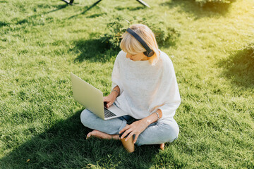 Mature adult Woman in headphones using laptop, sitting on the grass outside in park. Happy and smiling senior typing at notebook with coffee cup. Distance learning online education and work.