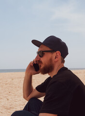 Bearded millennial man in black t-shirt snapback on blue sea landscape with smartphone in hand talking. Young adult guy calling friends on cell phone, real people communication lifestyle distance work