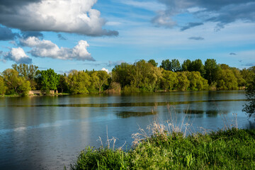Lakeshore looking across the lake to the opposite bank with trees under a blue sky with developing storm clouds.