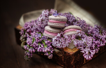 Obraz na płótnie Canvas Macaroni cakes lying on a bouquet of lilacs in a wooden box on a dark wooden background.