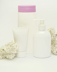 White cosmetic bottles with lilac on background. Scrub, gel shower and cream on light background with lilac petals.