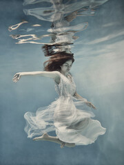A girl with long dark hair in a white dress with glitters floats underwater as if floating in zero...