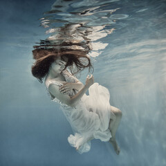 A girl with long dark hair in a white dress with glitters floats underwater as if floating in zero...