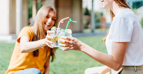 Two beautiful young women sitting on the grass and laughing while having a lemonade cocktails drink in glass jars during a friendly hang out in city park. Summer vacation and picnic at sunny day. - obrazy, fototapety, plakaty