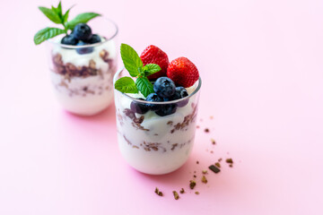 Joghurt mit Granola und frischen Beeren in zwei Gläser geschichtet. Gesundes Frühstück, servierfertig, pinker Hintergrund.