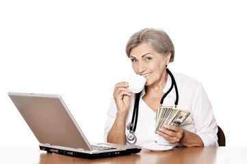 Senior doctor sitting at table with laptop and holding dollars