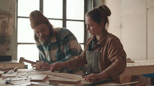 Medium shot of young female novice joiner standing at workbench in joinery workshop and watching bearded Caucasian man carving plank using gouge and small wooden hammer