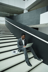 Graphic full length portrait of African-American businessman using laptop while sitting on stairs in office hall
