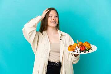 Teenager girl holding waffles over isolated blue background smiling a lot