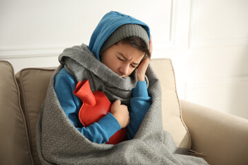 Sick teenage boy wrapped in blanket with hot water bottle on sofa at home