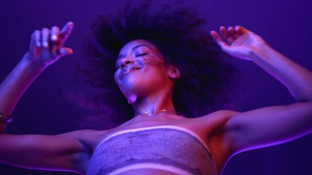 Amazing positive afro woman with curly hair dancing in nightclub under UV neon lamps. Charming lady, night life concept. Modern pop outfit - bandeau. High quality 4k footage