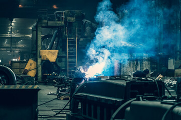 Welder in protective mask works in metallurgical factory workshop, sparks and smoke from welding flares, industrial background.