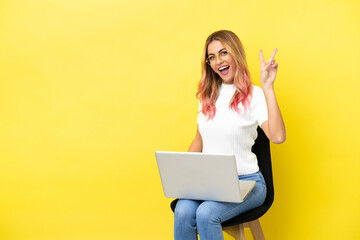 Young woman sitting on a chair with laptop over isolated yellow background smiling and showing victory sign