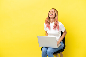 Young woman sitting on a chair with laptop over isolated yellow background laughing