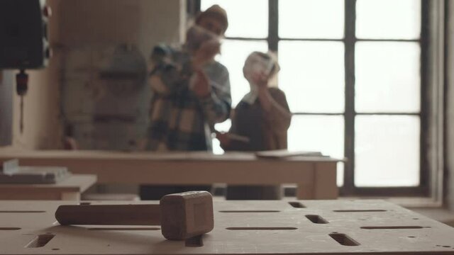 Lockdown of wooden hammer lying on workbench on foreground while male and female joiners working together on background in workshop