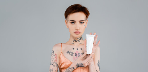 tattooed young woman holding tube with cosmetic product isolated on grey, banner