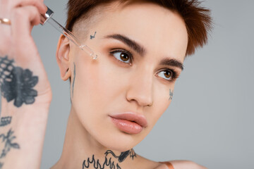 young tattooed woman applying serum with pipette isolated on grey