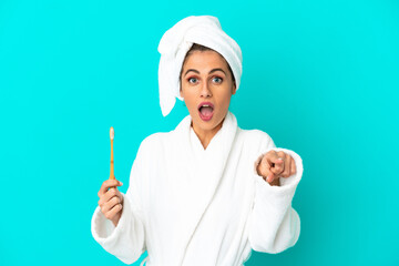 Young woman in a bathrobe brushing teeth over isolated blue background surprised and pointing front