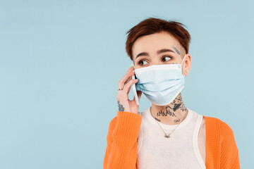 tattooed woman in orange cardigan and medical mask talking on smartphone isolated on blue