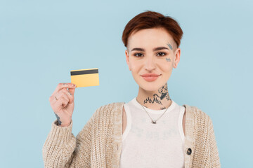 cheerful and tattooed woman holding credit card isolated on blue