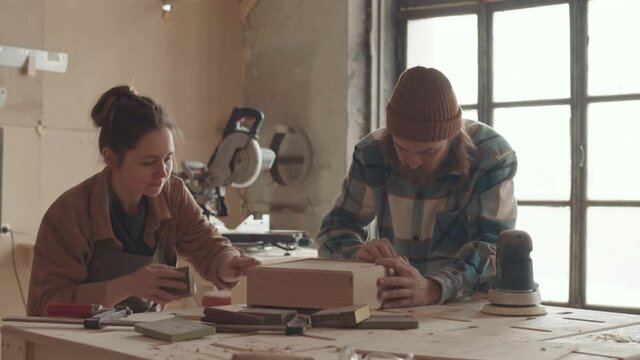 Medium shot of male and female Caucasian joiners standing at workbench in workshop and polishing wooden drawer together using sandpaper