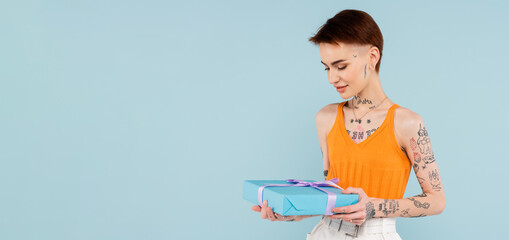 young tattooed woman looking at wrapped present isolated on blue, banner