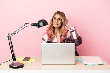 Young student woman in a workplace with a laptop over pink background having doubts
