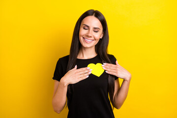 Photo of adorable satisfied girl closed eyes hands hold small paper heart postcard isolated on yellow color background