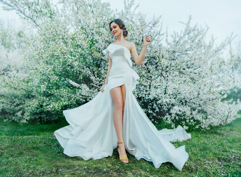 Fantasy woman in long white elegant fashion long dress walks in green spring blossom cherry garden. Happy cheerful girl princess bride Skirt fabric flies flowing waving in wind motion. Sexy bare legs