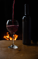 Wine, wine bottle and glass being filled with wine on rustic wooden surface with fire in the background, low key image, dark background, selective focus.