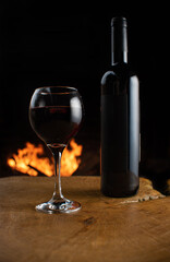 Wine, full wine glass and wine bottle on rustic wooden surface with fire in the background, low key image, dark background, selective focus.