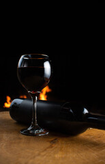 Naklejka na ściany i meble Wine, full wine glass and wine bottle lying on rustic wooden surface with fire in the background, low key image, dark background, selective focus.