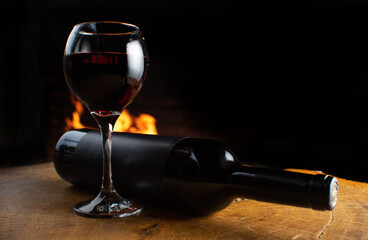 Wine, full wine glass and wine bottle lying on rustic wooden surface with fire in the background, low key image, dark background, selective focus.