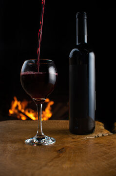 Wine, wine bottle and glass being filled with wine on rustic wooden surface with fire in the background, low key image, dark background, selective focus.