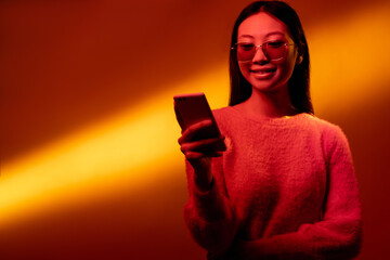 Digital people. Mobile technology. Color light portrait. Online communication. Neon pink smiling Asian girl using phone isolated on yellow golden gradient copy space background.