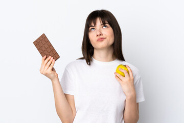 Young Ukrainian woman isolated on white background having doubts while taking a chocolate tablet in one hand and an apple in the other