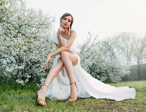 Happy woman fashion model sitting posing. long sexy bare legs white evening wedding dress. Romantic lady Adult girl bride in perfect gown. Nature background green blooming trees. cherry flowers garden